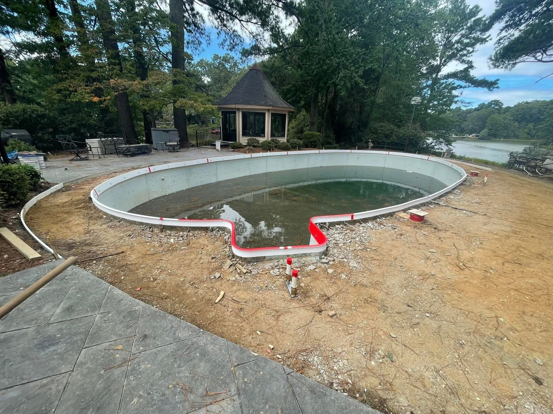 Empty, oval-shaped swimming pool under construction outdoors, surrounded by dirt, with a gazebo in the background.