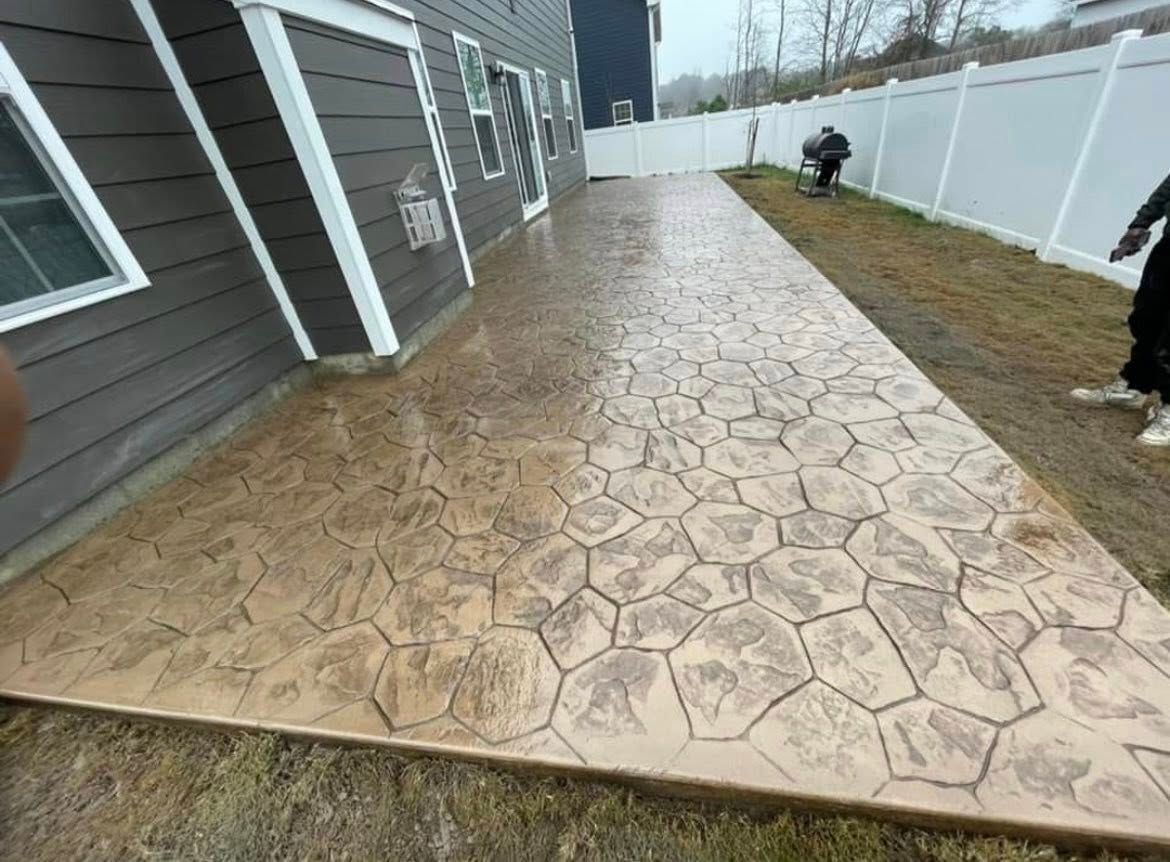 Stamped concrete patio, brown stone pattern, next to a gray house and white fence.