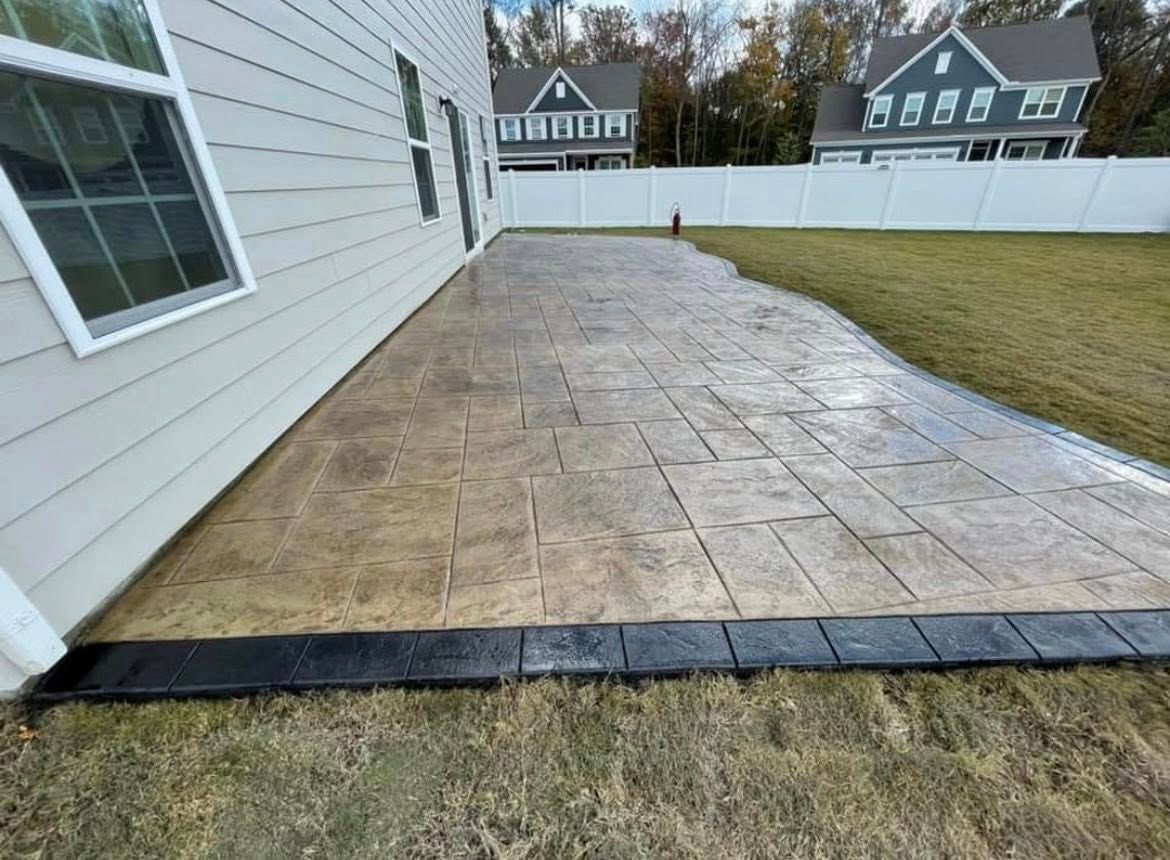 Stamped concrete patio adjacent to a light gray house. Bordered by black trim and lawn.