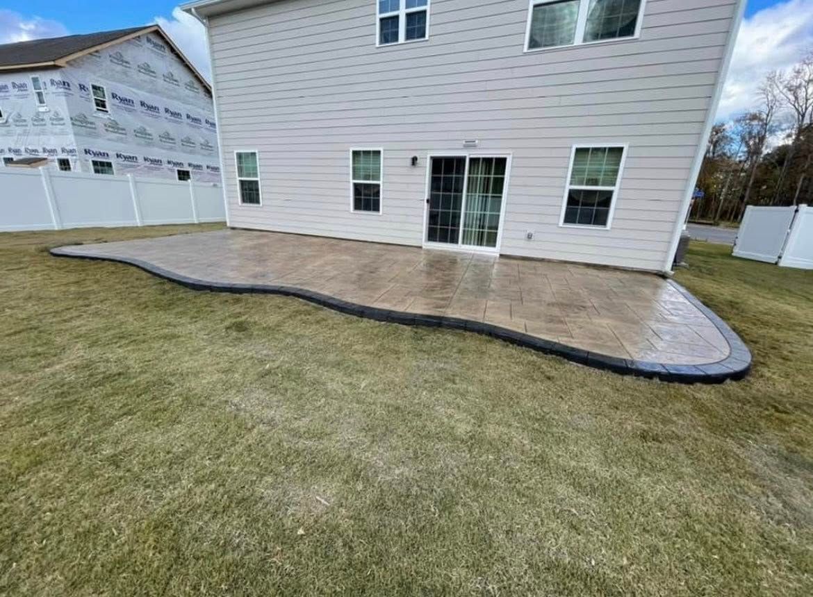 Backyard patio with a dark border, adjacent to a two-story beige house and grassy lawn.