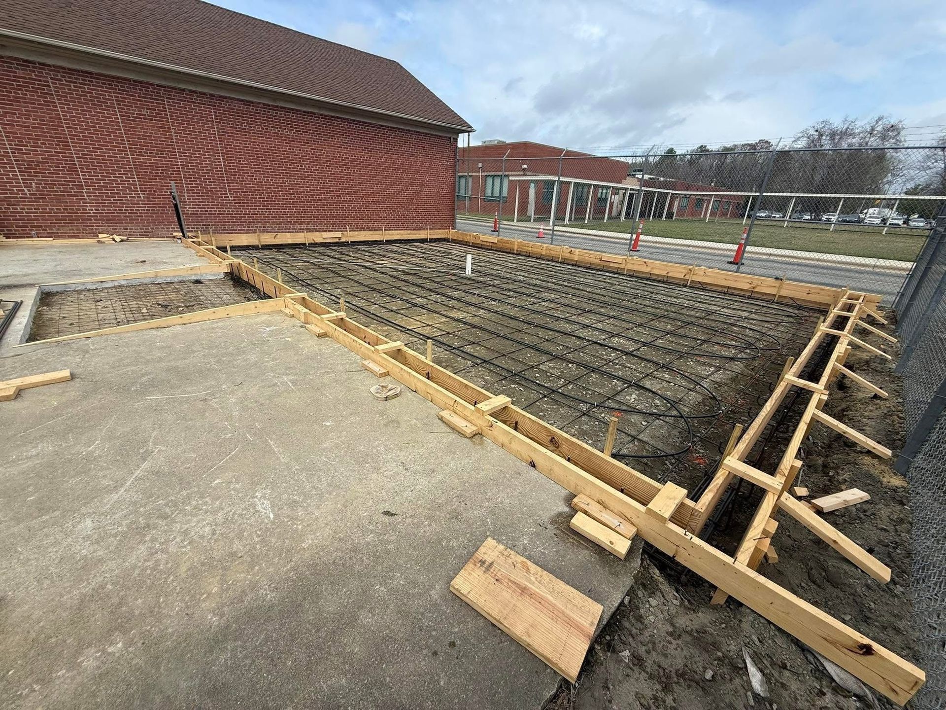 Construction site: Wooden forms define a concrete pad with rebar reinforcement; next to a building and paved area.