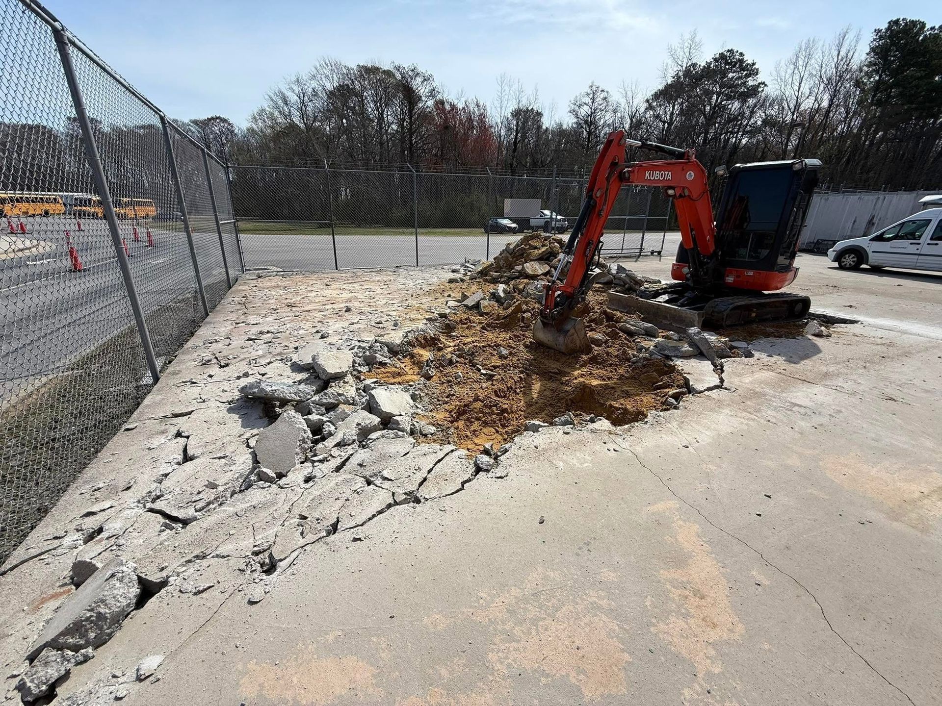 Mini excavator breaking up concrete next to a chain-link fence.