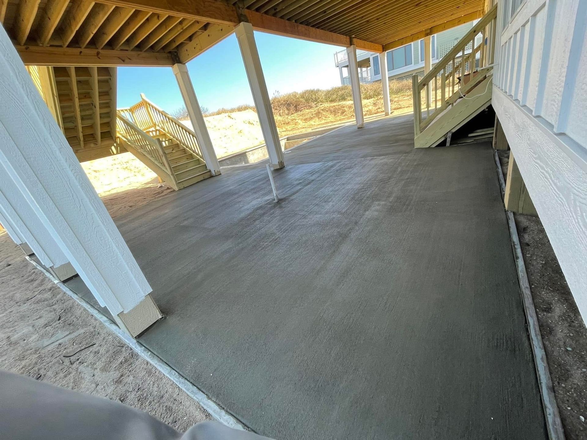 Newly poured concrete patio under a raised deck, with wooden supports and stairs, on a sunny day.