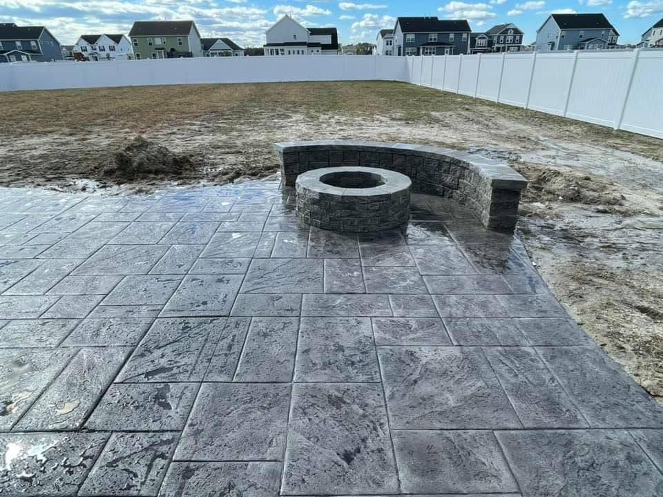 Concrete patio with built-in fire pit and curved seating in a backyard with a white fence.