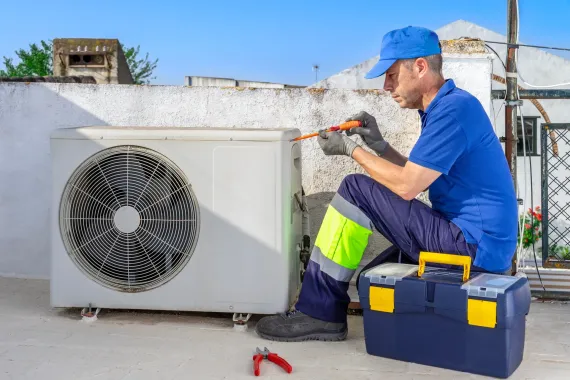 HVAC technician repairs an air conditioning unit outdoors, wearing blue and neon work gear, holding a screwdriver.