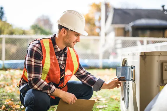 HVAC technician in hard hat and safety vest inspects outdoor unit, kneeling in grass.