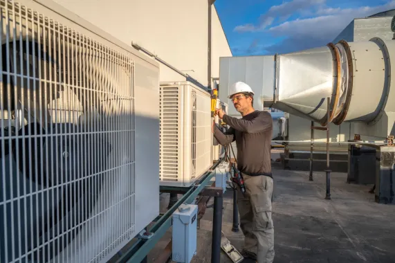 HVAC technician in white hard hat repairs rooftop air conditioning unit.