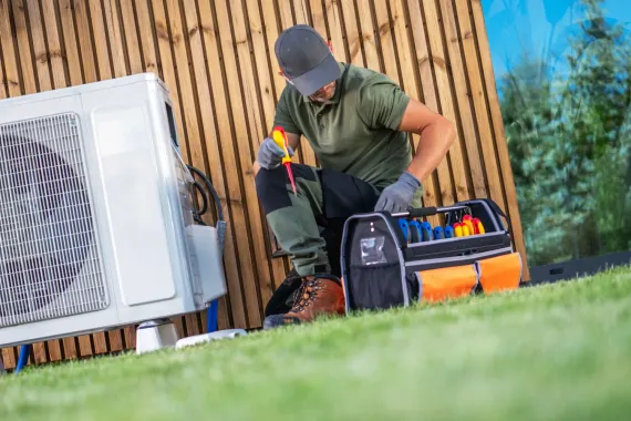 HVAC technician repairs air conditioner unit outdoors, wearing gloves, cap, and work clothes. Tool bag nearby.