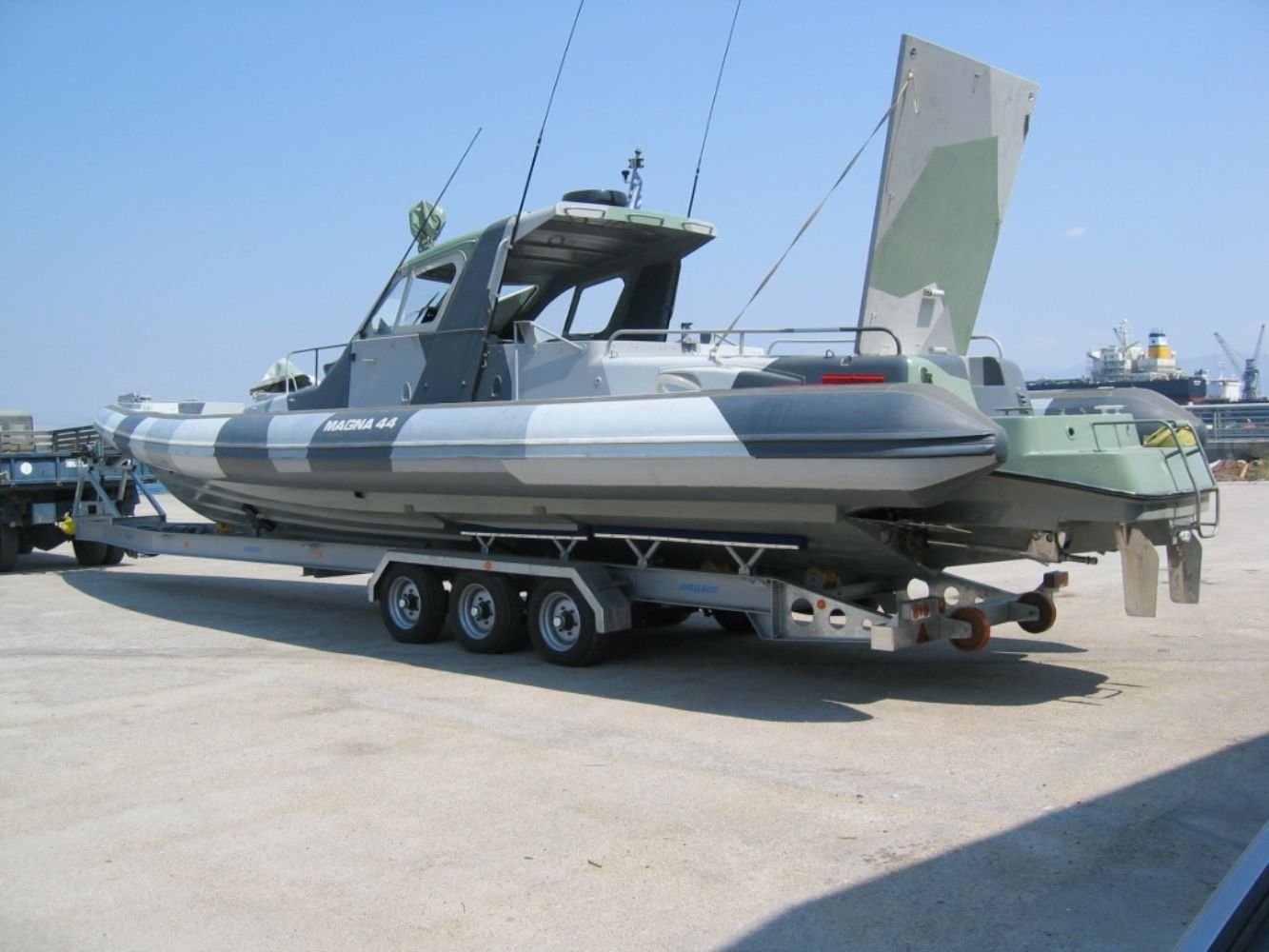 Imbarcazione militare mimetizzata su un rimorchio, parcheggiata su una superficie di cemento. Cielo azzurro sullo sfondo.