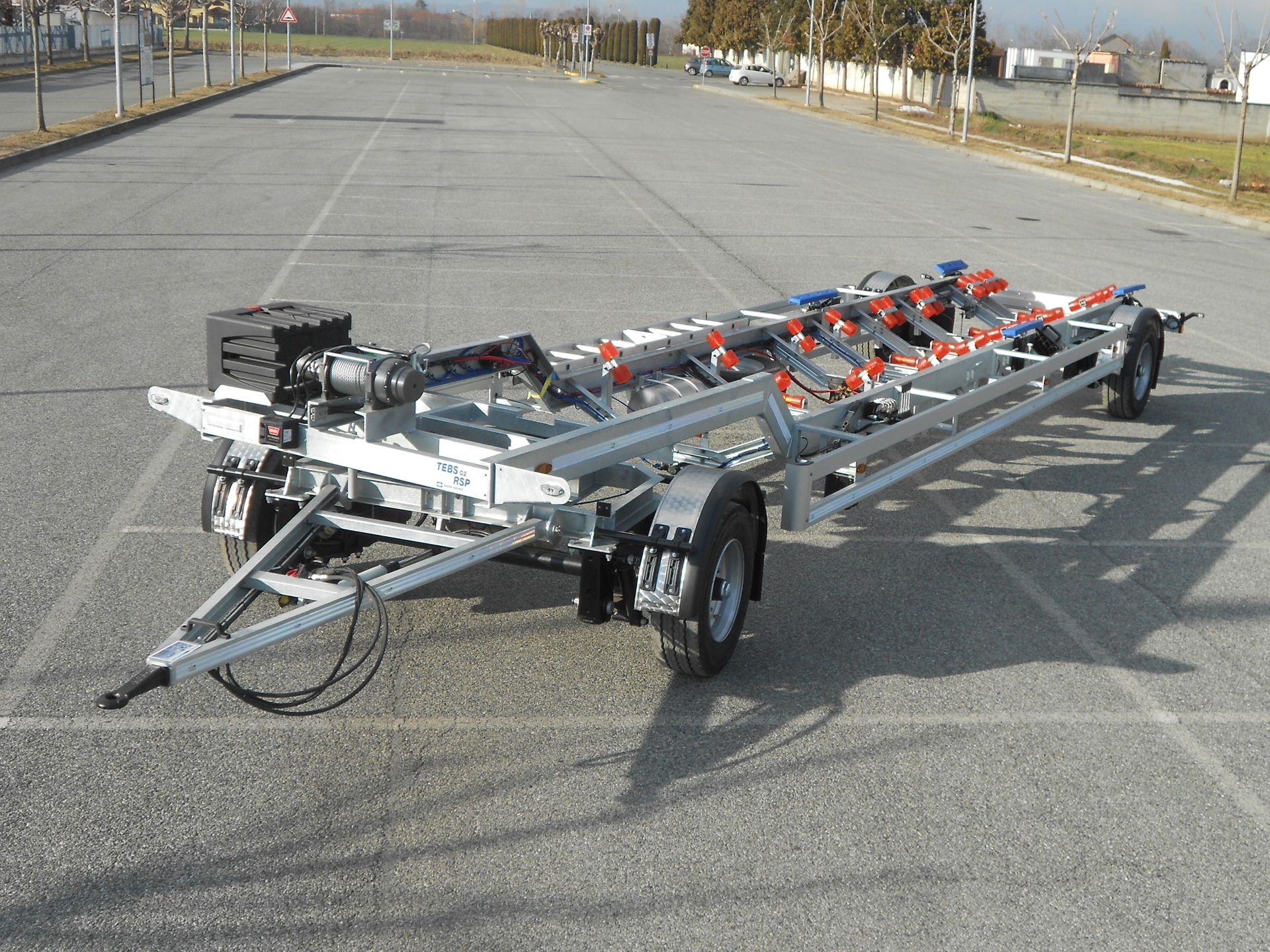 Rimorchio per barche parcheggiato su un terreno asfaltato; telaio in acciaio con rulli e verricello.