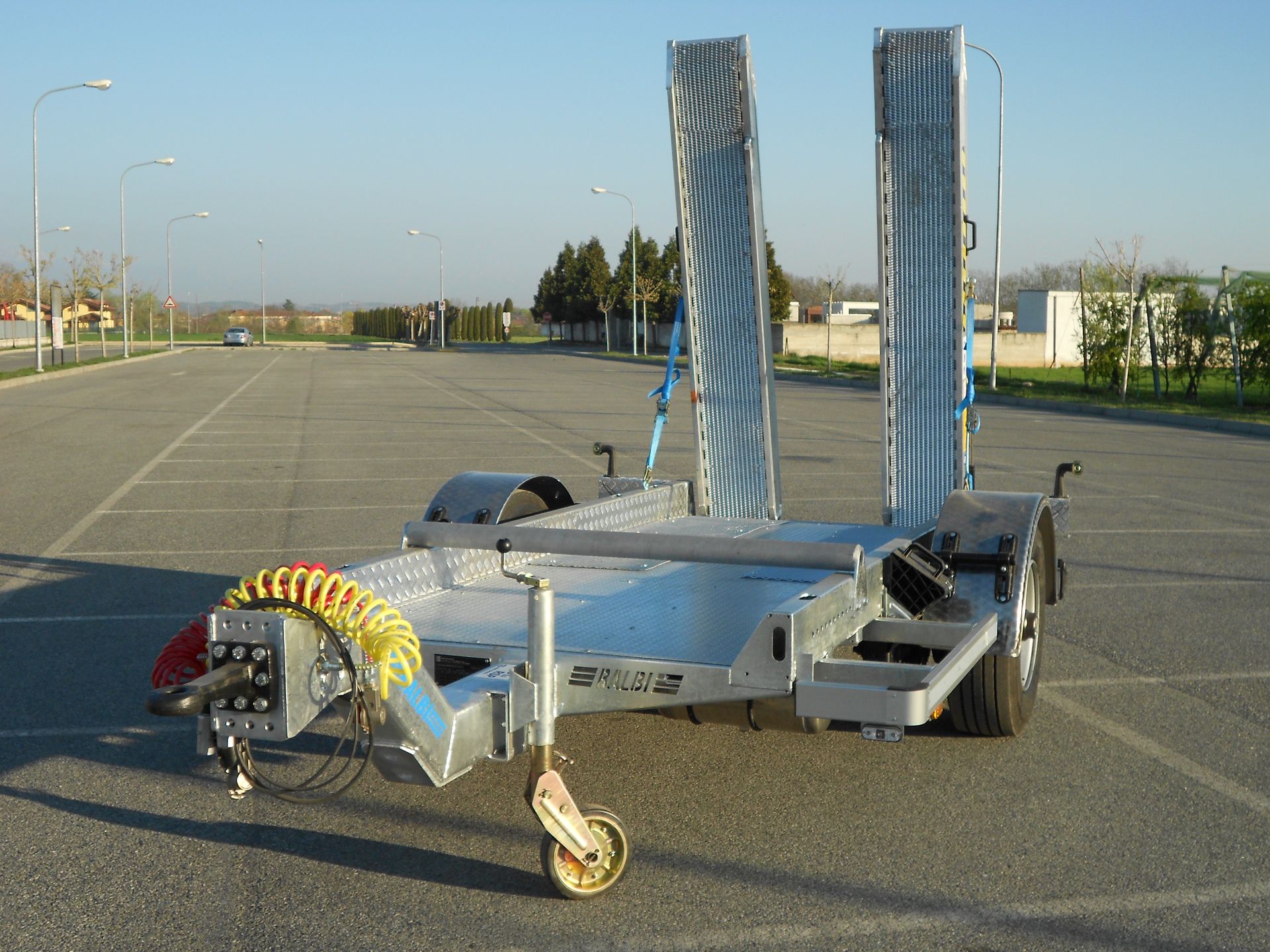 Rimorchio per auto color argento con due rampe, parcheggiato su un piazzale asfaltato.