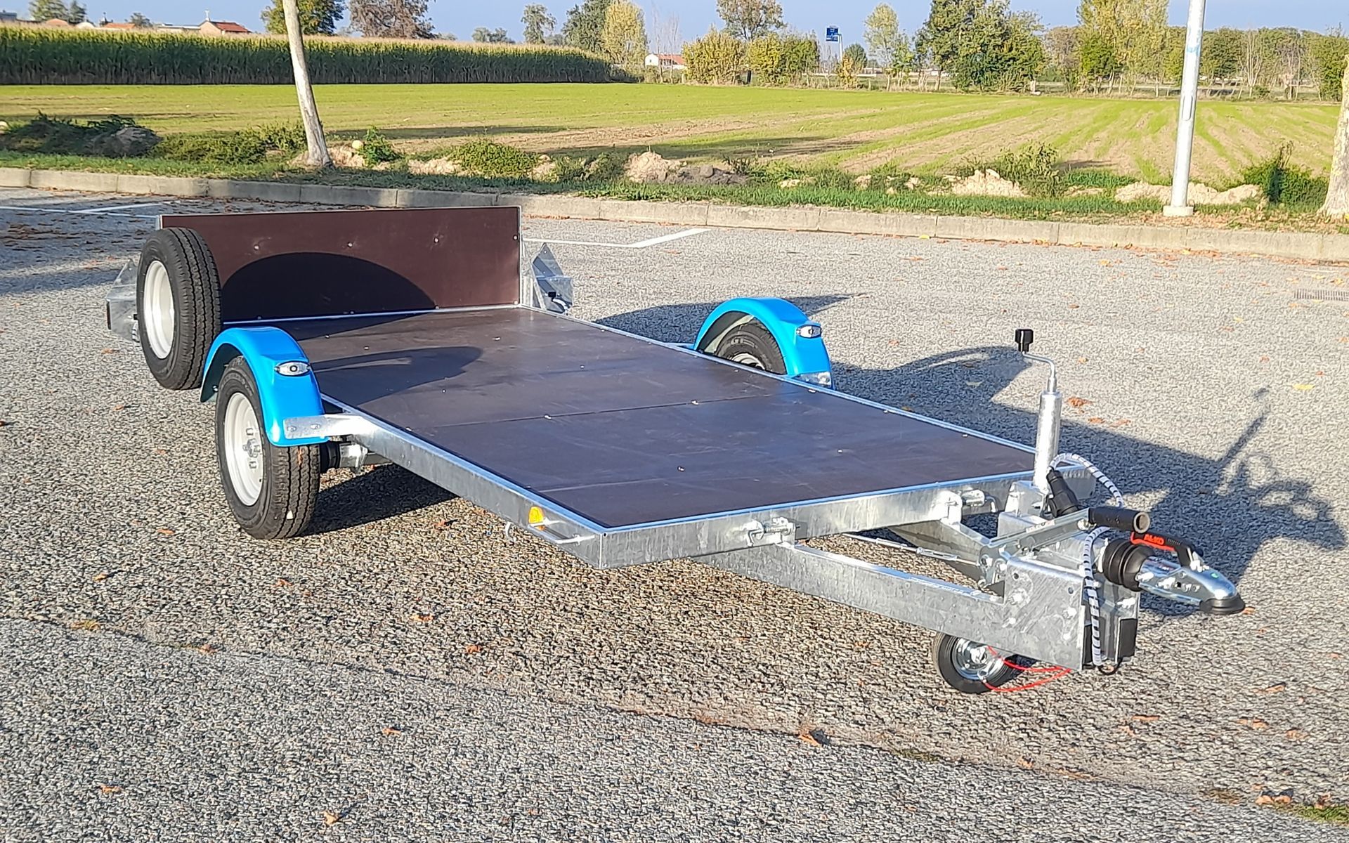 Rimorchio utilitario con pianale, parafanghi blu, ruota di scorta e sfondo di ghiaia.