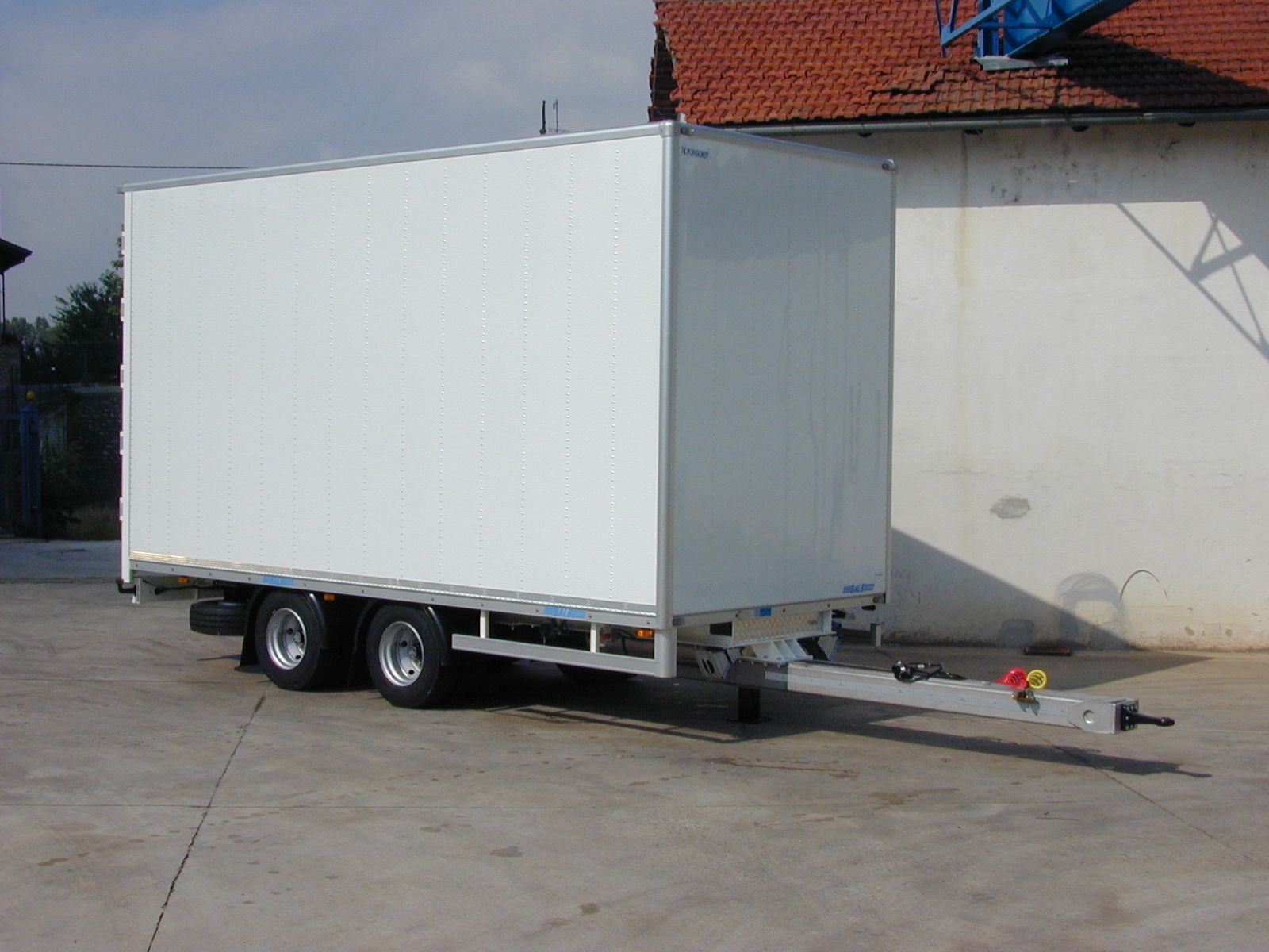 Rimorchio bianco su ruote, parcheggiato sul cemento di fronte a un edificio con tetto di tegole rosse.