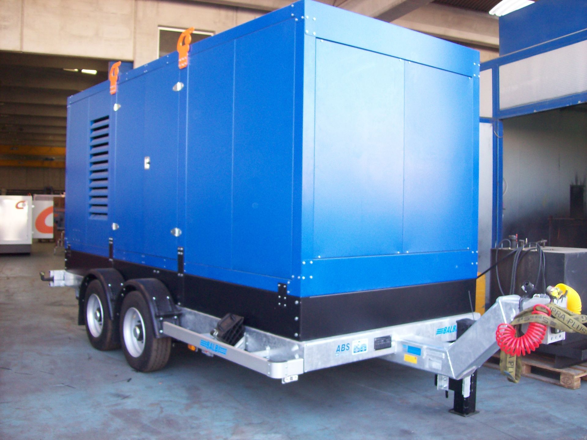 Generatore blu su un rimorchio, con due assi, in un ambiente industriale.