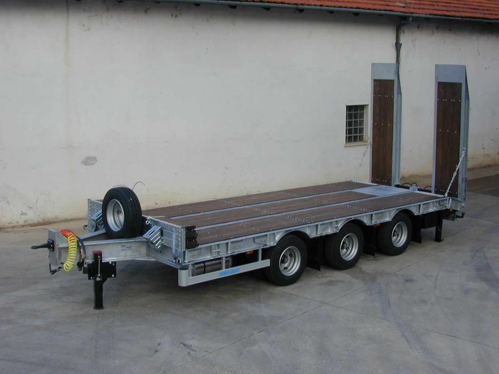 Rimorchio ribassato con quattro assi e due rampe, parcheggiato all'aperto davanti a un edificio.