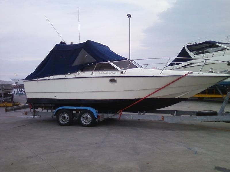 Motoscafo bianco su rimorchio, coperto da un telo blu, parcheggiato all'aperto.