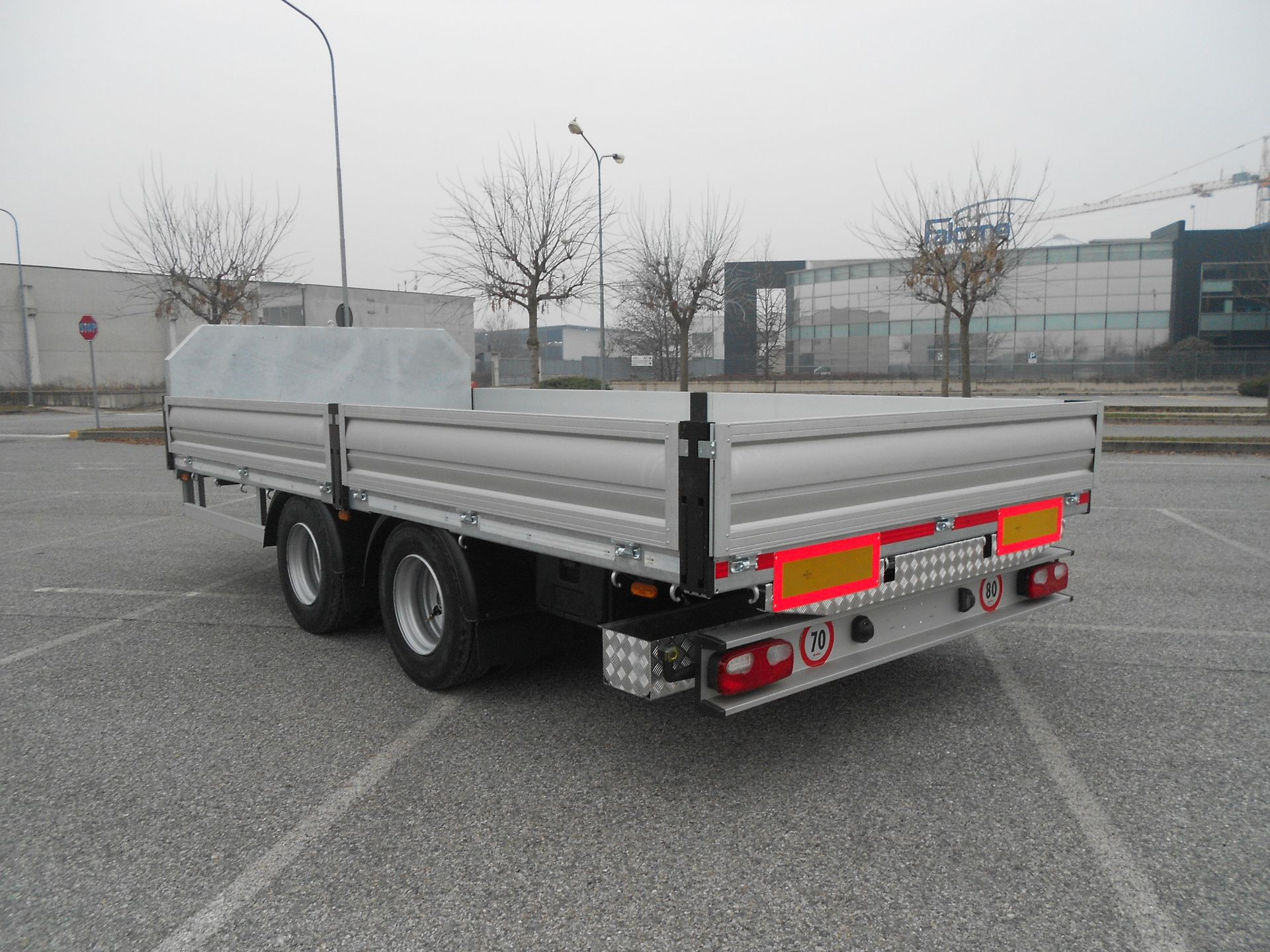 Vista posteriore di un rimorchio a pianale argentato con tre assi parcheggiato sull'asfalto.