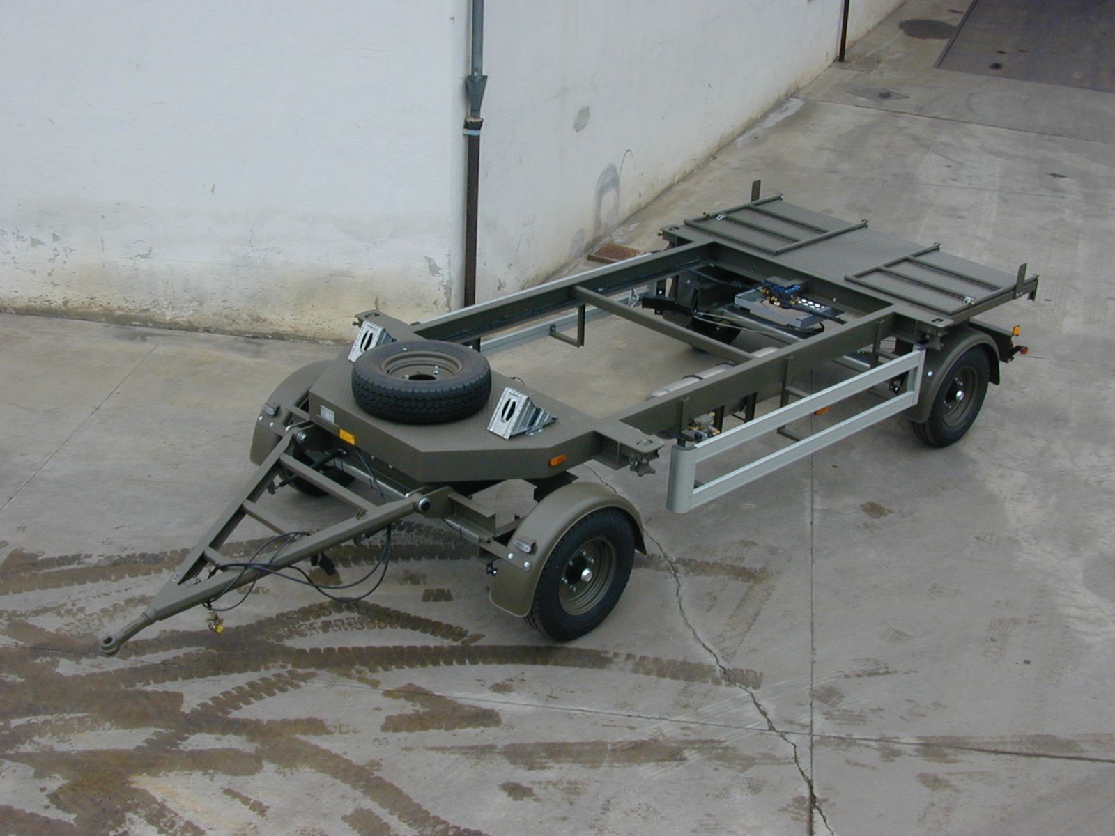 Rimorchio verde militare con ruota di scorta, gancio di traino e due assi su una superficie di cemento.
