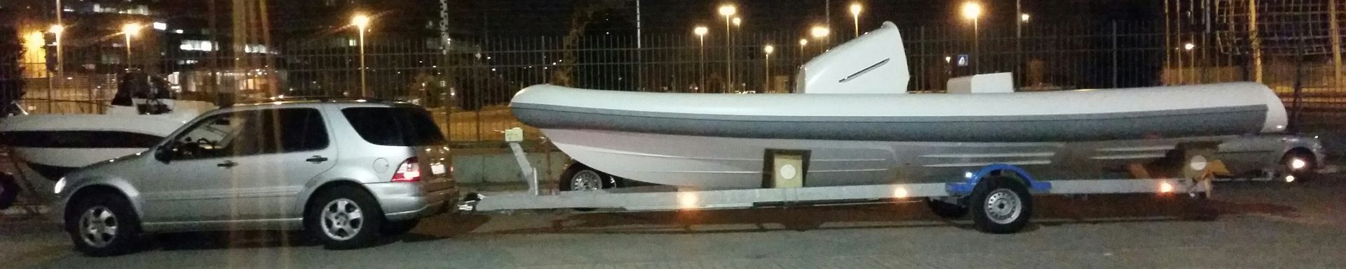 Un SUV argentato che traina un grande gommone rigido (RIB) bianco su un rimorchio, parcheggiato di notte.