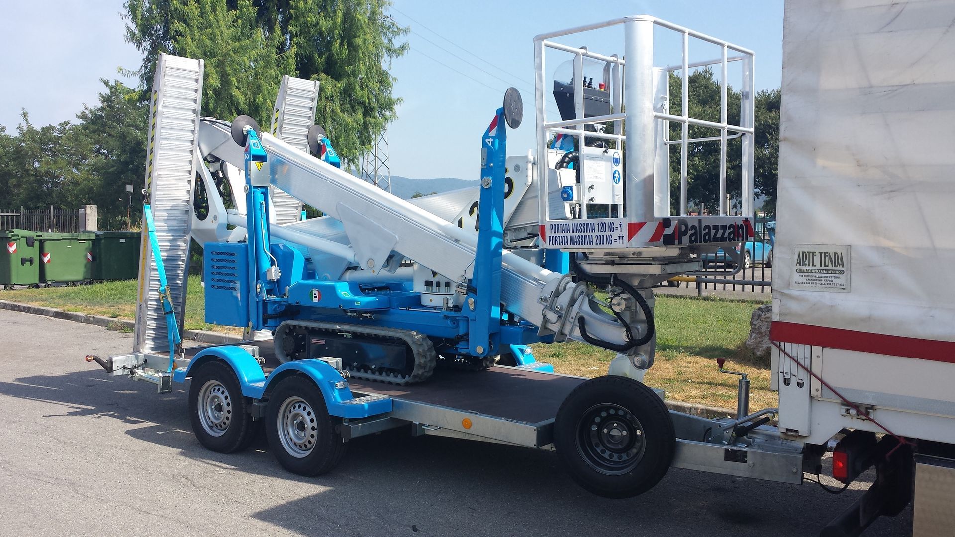 Un ascensore blu su un rimorchio, parcheggiato accanto a un camion bianco in una giornata di sole.