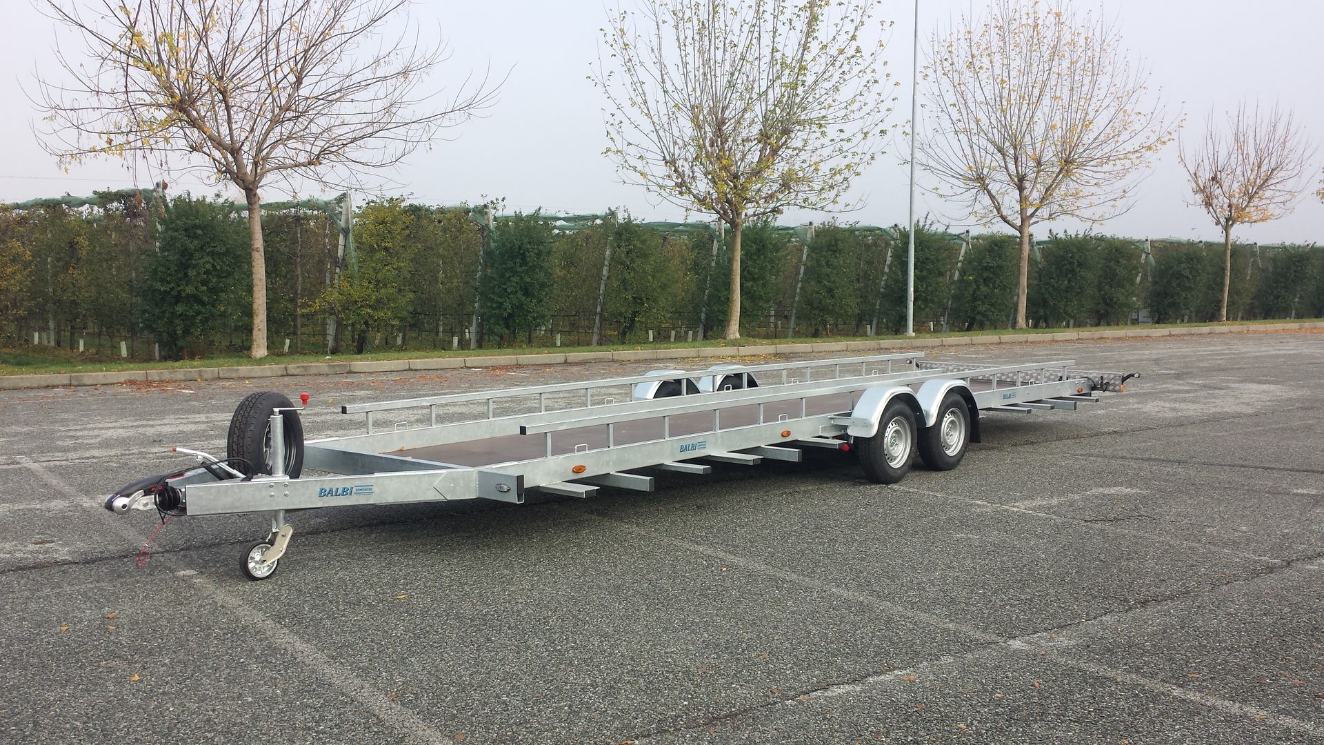 Rimorchio per barche vuoto su un parcheggio asfaltato, alberi sullo sfondo, giornata nuvolosa.