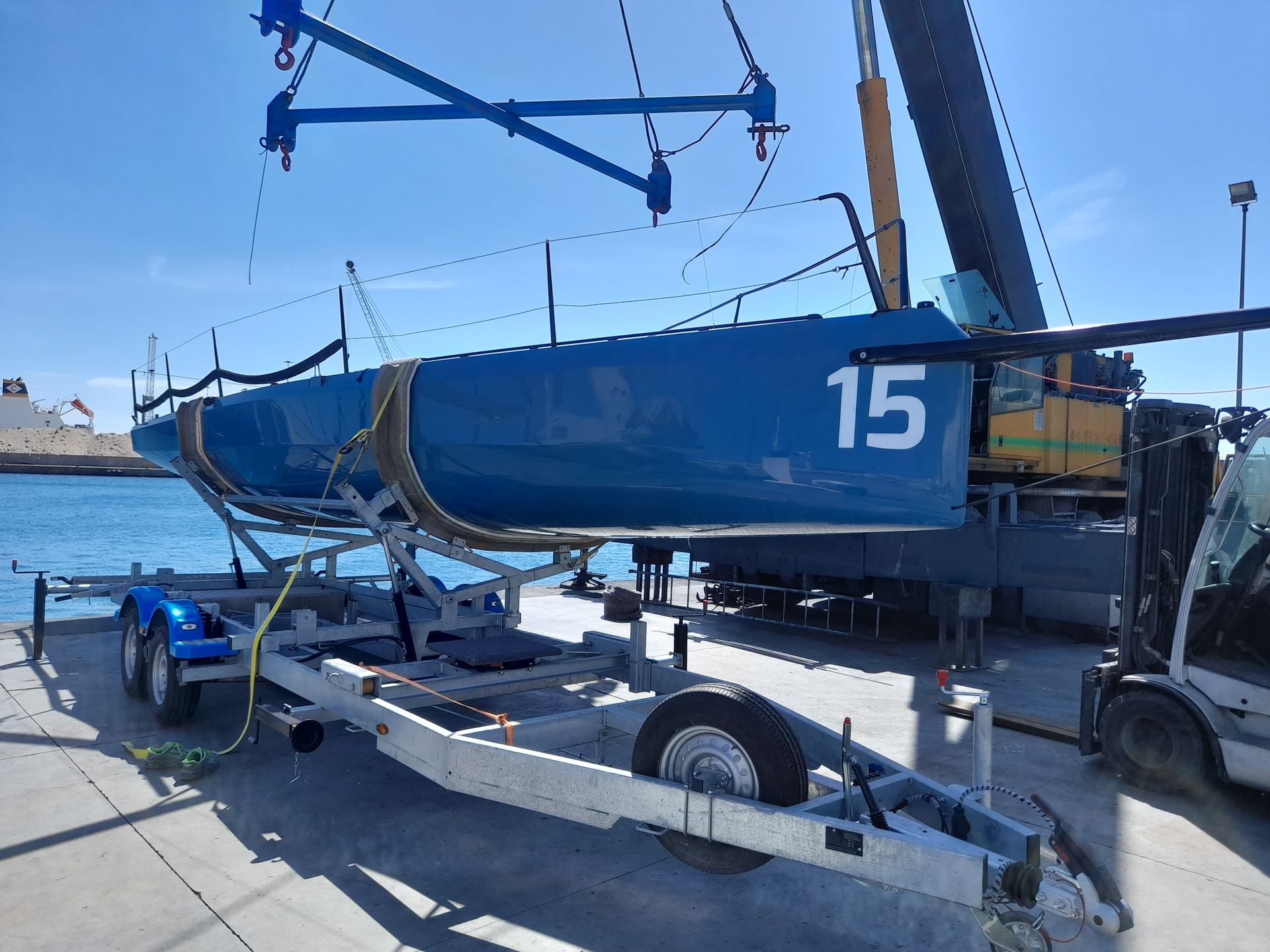 Barca a vela blu su un rimorchio, sollevata da una gru vicino a un porto.