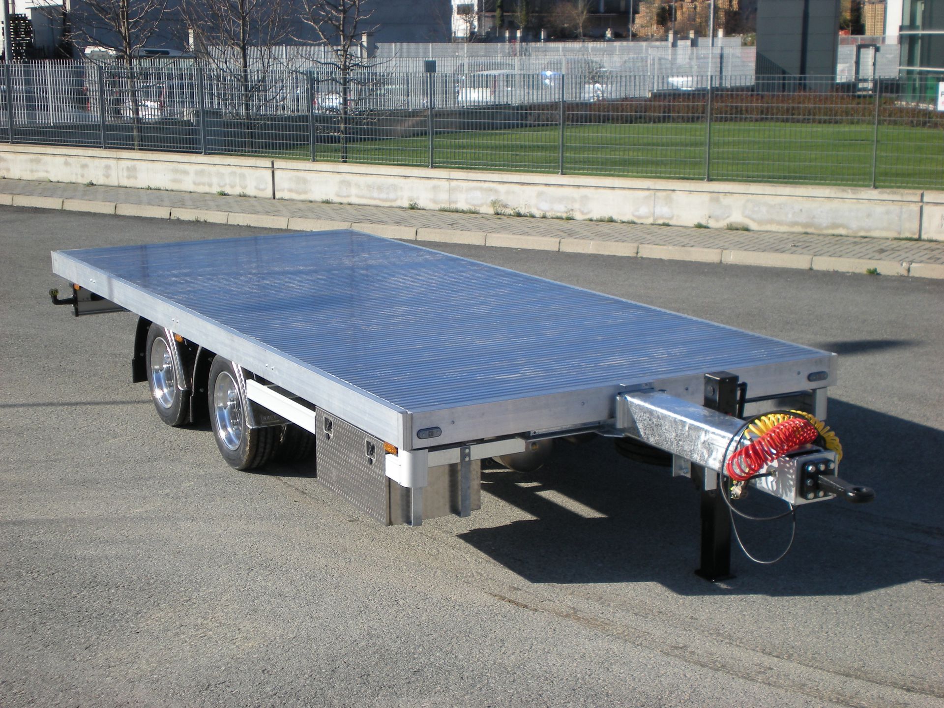 Rimorchio con pianale su asfalto, con pianale in lamiera ondulata color argento e gancio di traino nero.