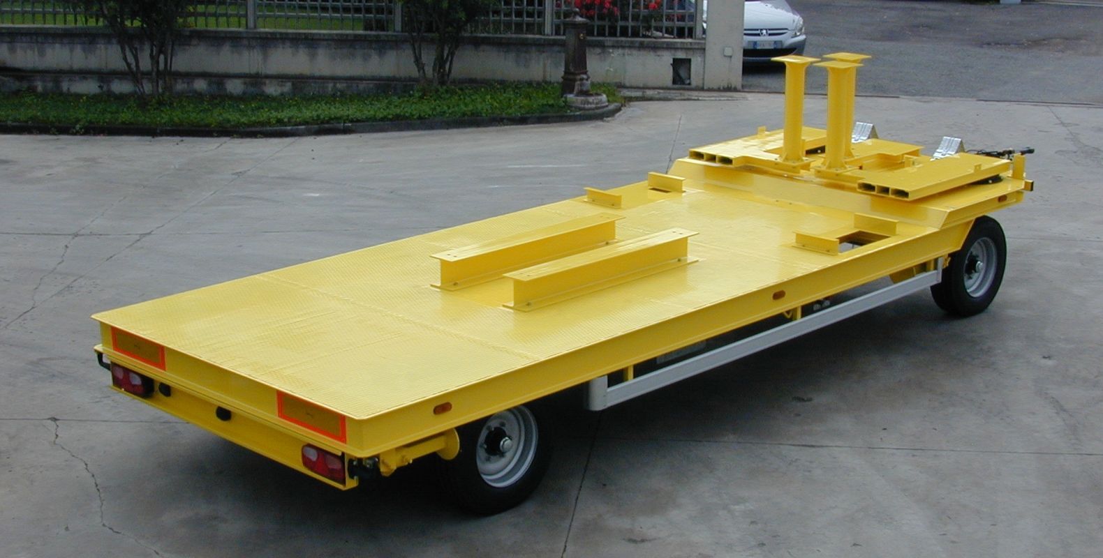 Rimorchio industriale giallo con pianale, pneumatici e supporti verticali su una superficie di cemento.
