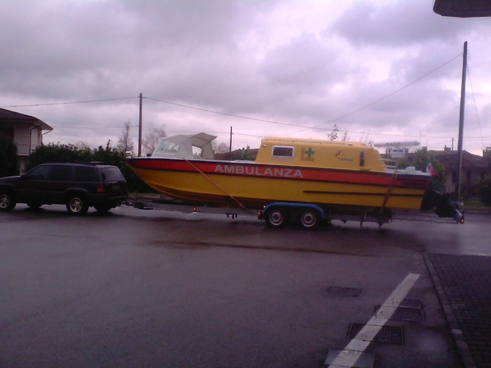 Un SUV nero che traina un'ambulanza gialla su un rimorchio, parcheggiato su una strada bagnata sotto un cielo nuvoloso.
