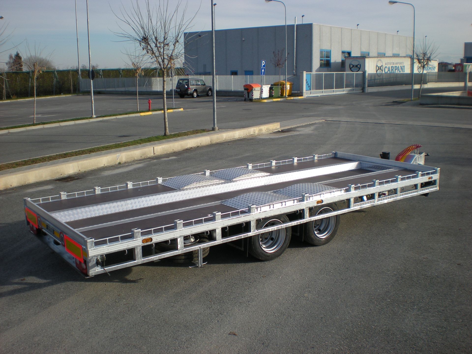 Un rimorchio a pianale parcheggiato sull'asfalto in un parcheggio. È argento e nero, con tre assi.