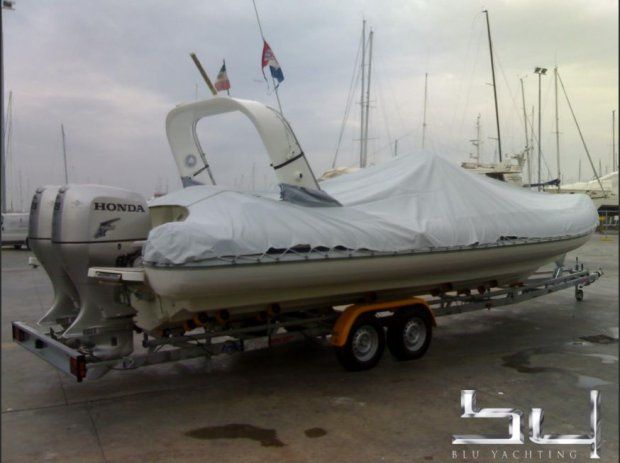 Un gommone rigido bianco con telo di copertura su un rimorchio, con due motori fuoribordo Honda, in un porto turistico.