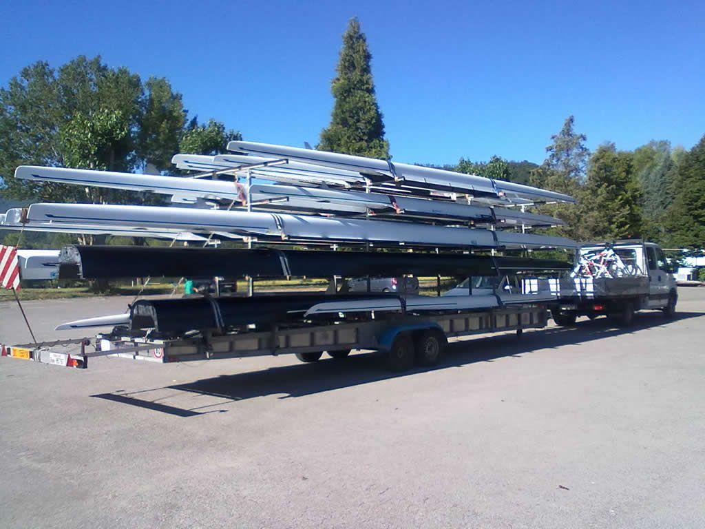 Un camion che traina un rimorchio carico di numerose barche da canottaggio argentate in una giornata di sole.