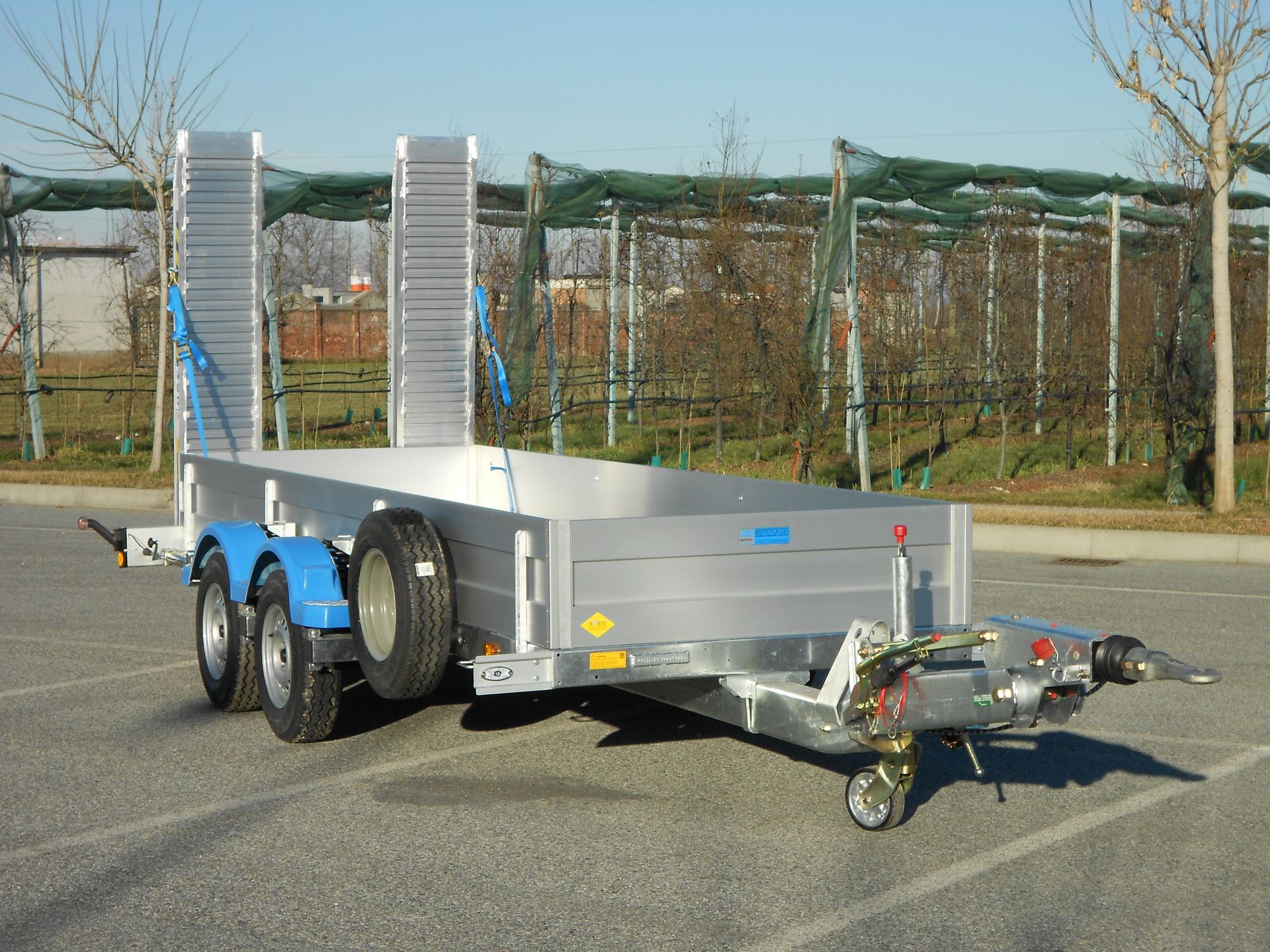 Rimorchio utilitario color argento con rampe, dettagli blu, ruota di scorta, parcheggiato sul marciapiede.