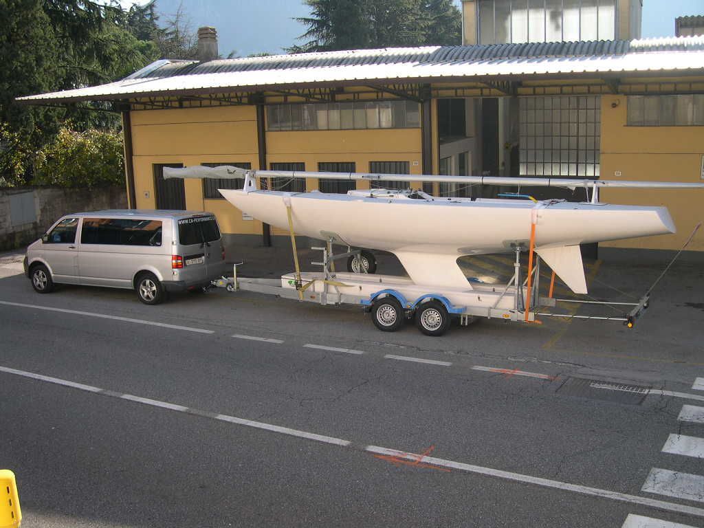 Una barca a vela bianca su un rimorchio viene trainata da un furgone argentato su una strada asfaltata.