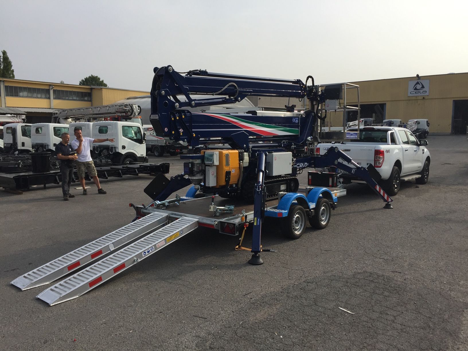 Una gru blu su un rimorchio agganciato a un pick-up bianco sull'asfalto. Le rampe sono abbassate.