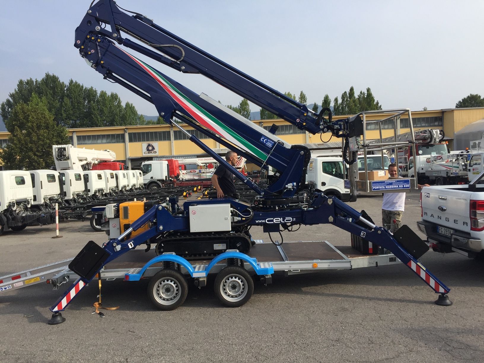 Gru articolata blu su rimorchio, con stabilizzatori estesi. All'aperto, vicino a un camion bianco.