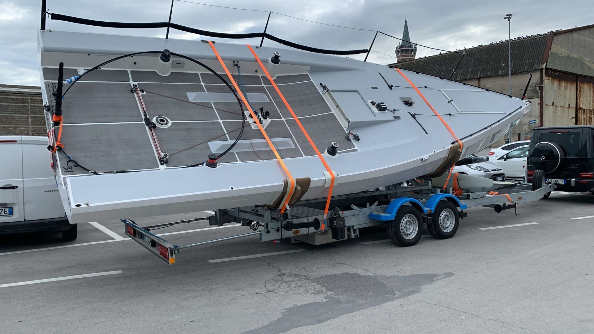 Barca a vela bianca su un rimorchio, assicurata con cinghie arancioni, parcheggiata su un terreno asfaltato.