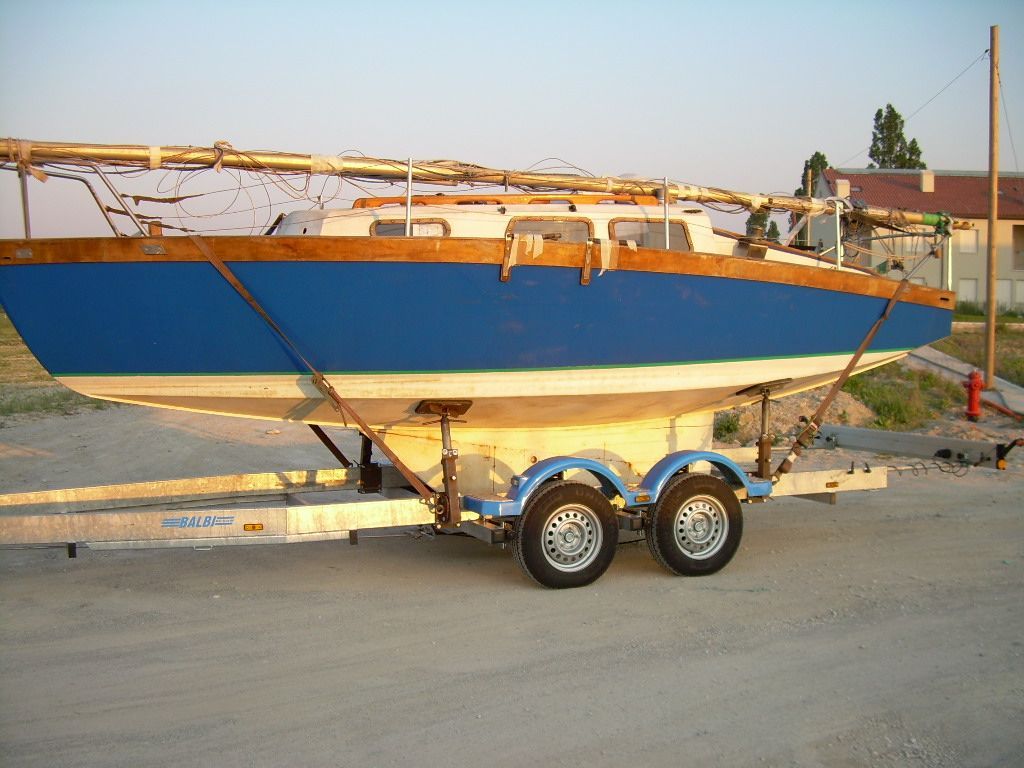 Barca a vela blu su rimorchio, con scafo bianco e finiture in legno, parcheggiata su un'area ghiaiosa.