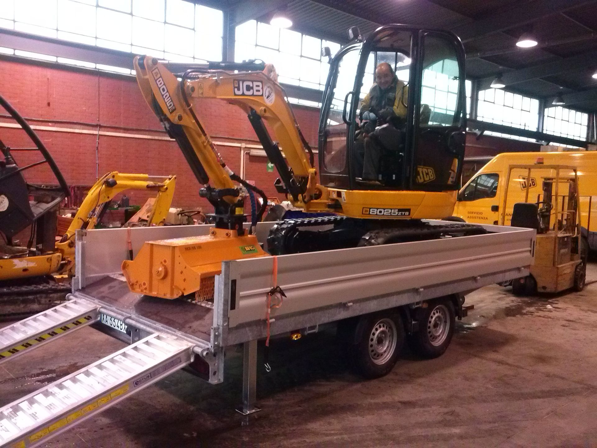 Escavatore JCB giallo su rimorchio con rampa, al chiuso con una persona in cabina.