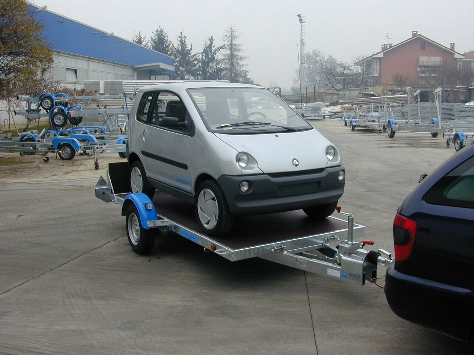 Un'auto argentata su un rimorchio, agganciata a un'auto blu, parcheggiata su un piazzale in cemento con rimorchi.