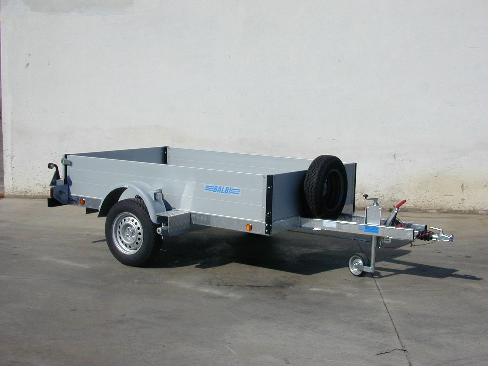 Rimorchio grigio con ruota di scorta contro un muro bianco.