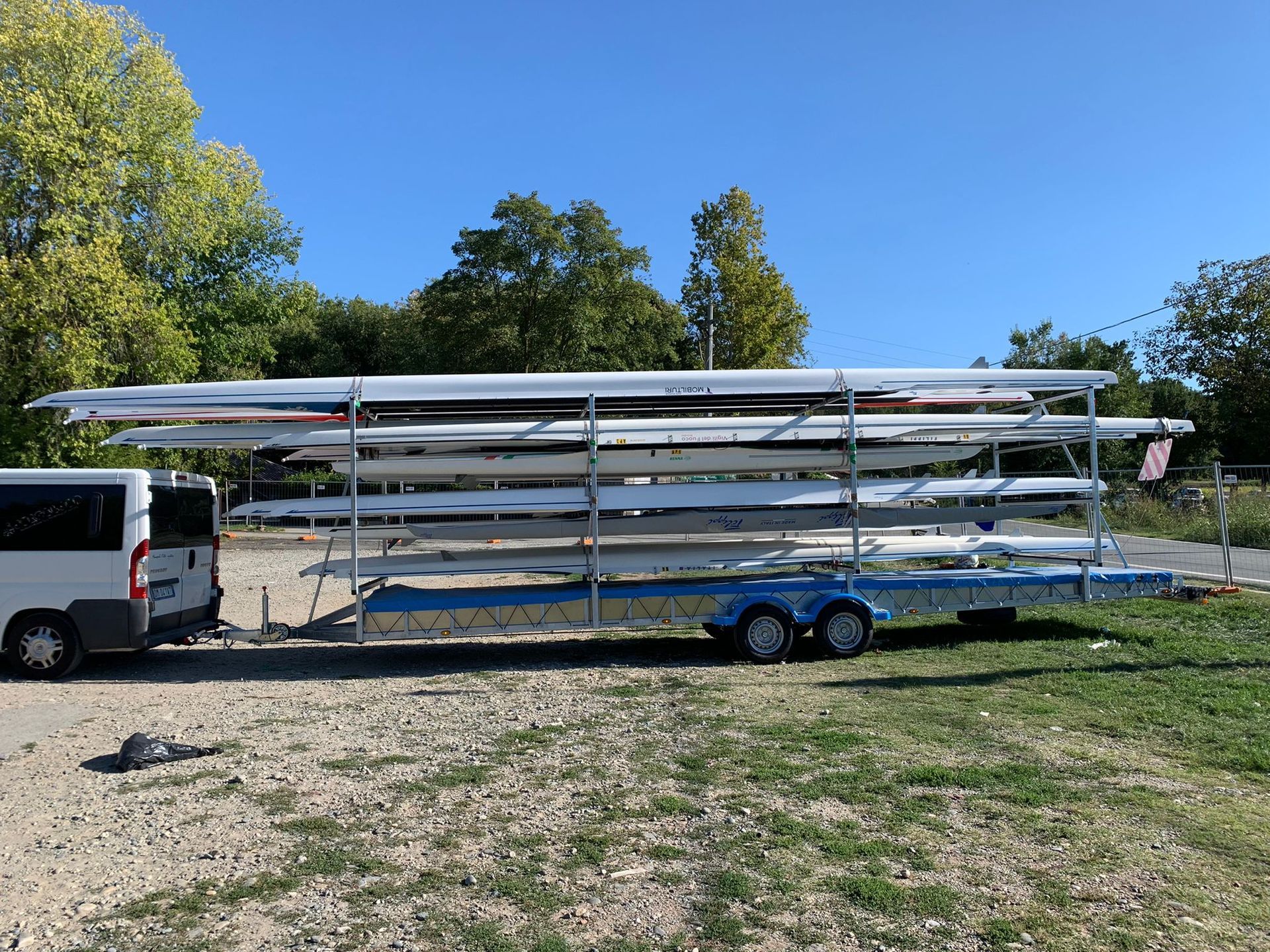 Un rimorchio carico di diverse lunghe barche da canottaggio bianche era parcheggiato sulla ghiaia accanto a un'area erbosa.