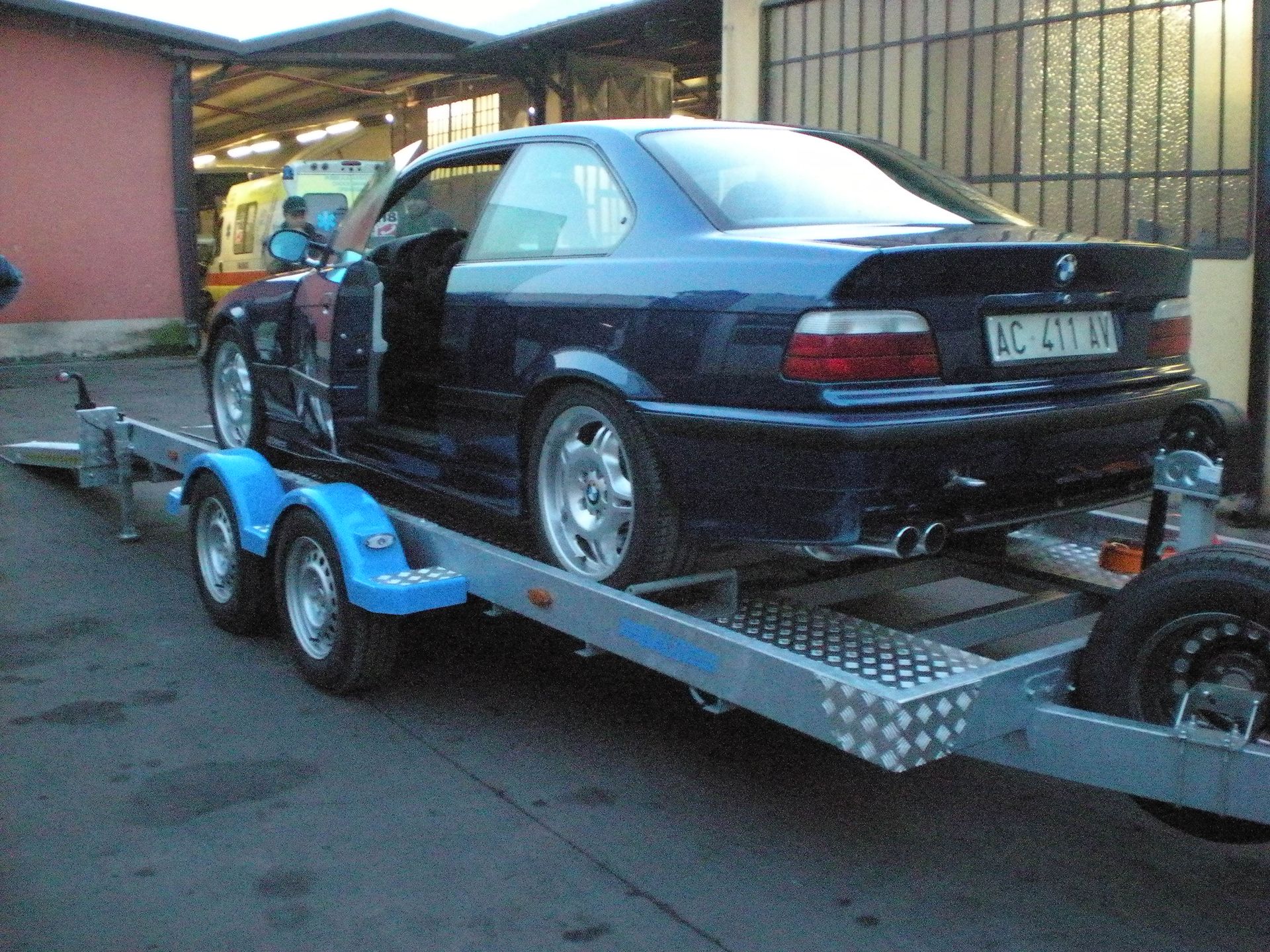 BMW coupé blu su rimorchio, portiera aperta. Esterno, ambientazione serale, trasporto veicoli.