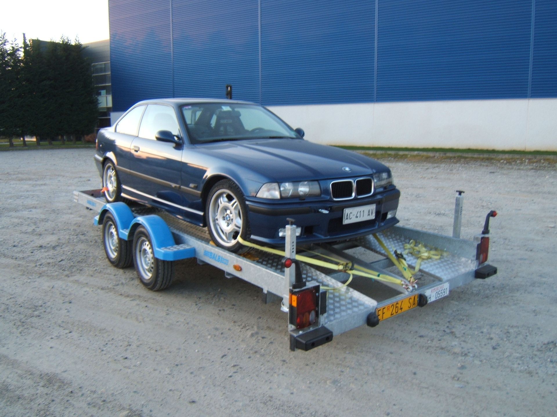Una BMW coupé blu su un rimorchio per auto, parcheggiata all'aperto, con un edificio blu sullo sfondo.