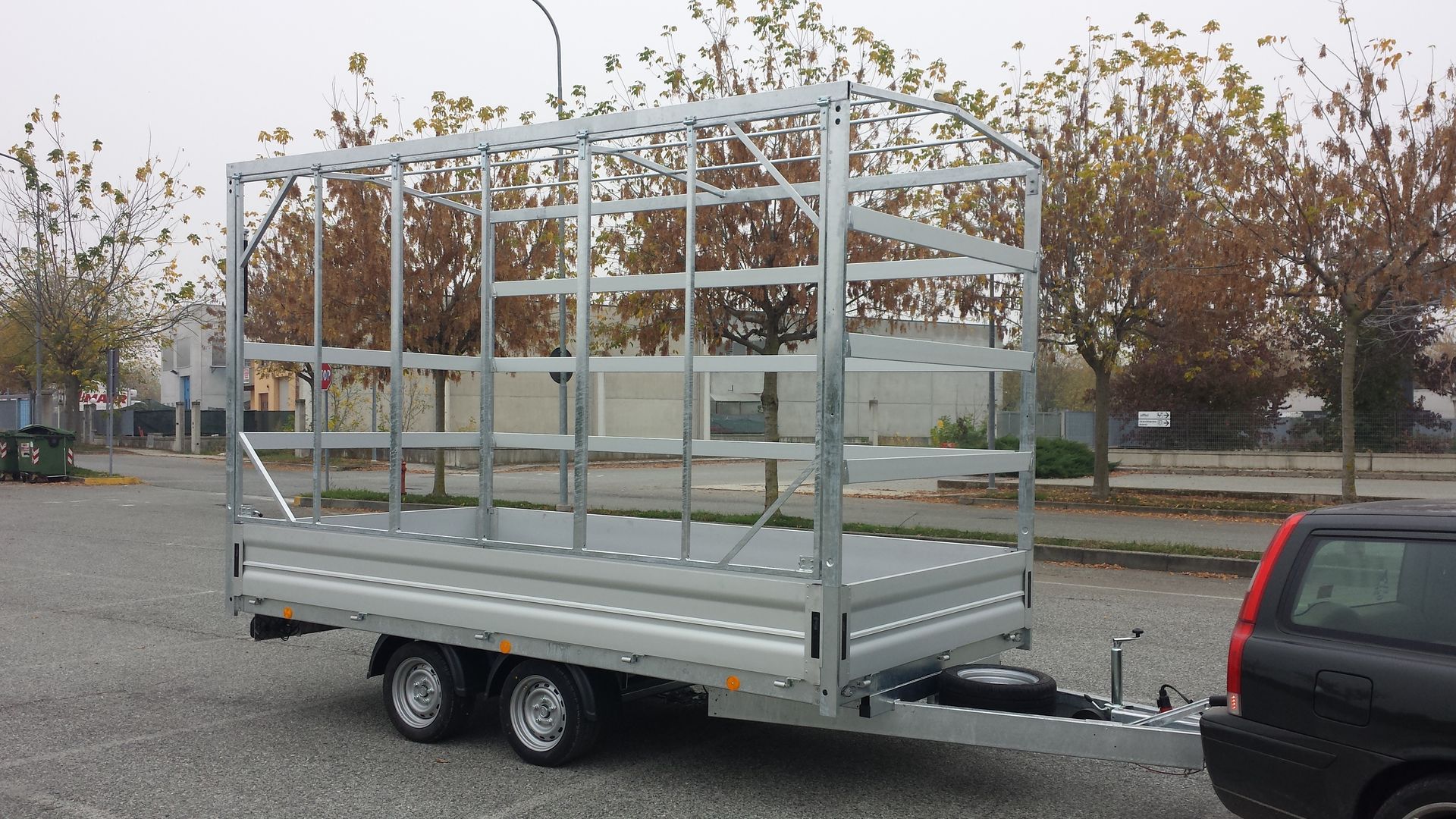 Un rimorchio grigio, scoperto, con una struttura metallica, parcheggiato all'aperto accanto a un'auto scura.