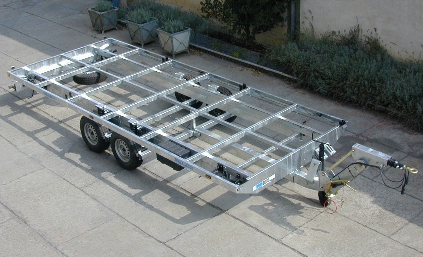 Telaio di rimorchio in metallo argentato su una superficie di cemento, con due assi e un gancio.