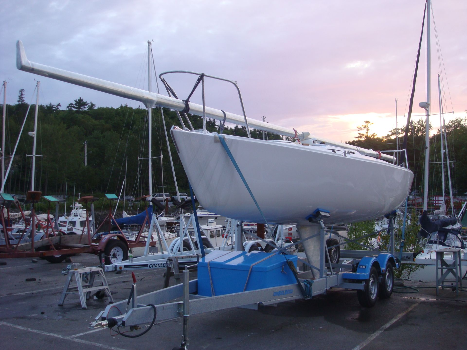 Barca a vela bianca su rimorchio, con albero esteso, in un porto turistico, cielo al tramonto.