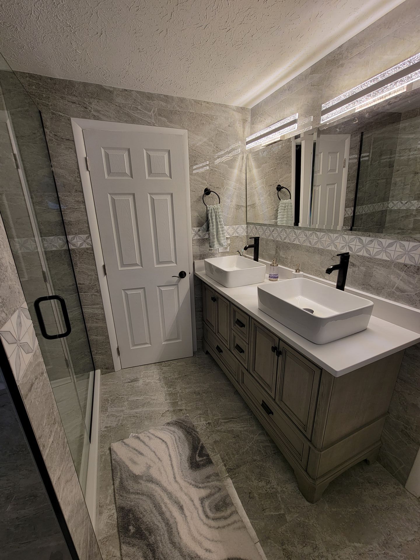 Bathroom with double vanity, white sinks, gray tile, white door, and glass shower.