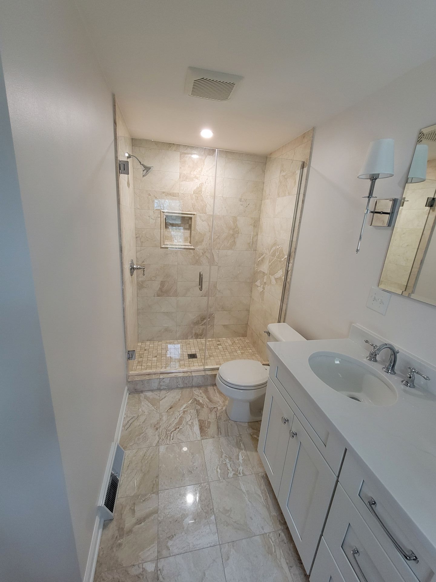 Small bathroom with a tiled shower, vanity, and toilet; light-colored stone and white fixtures.