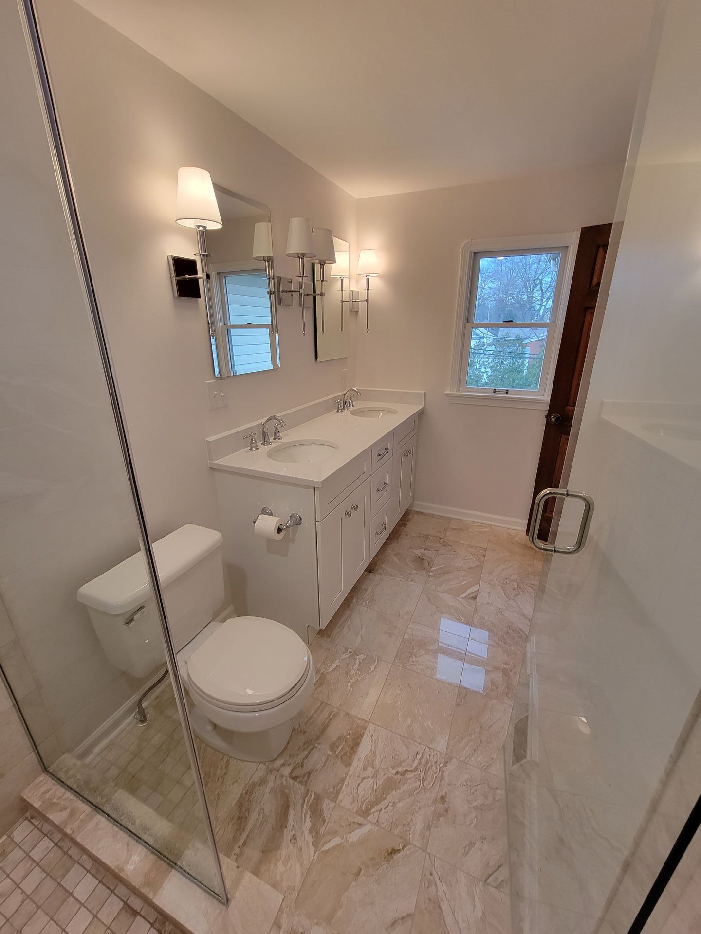 White bathroom with dual vanity, toilet, glass shower, and marble flooring.